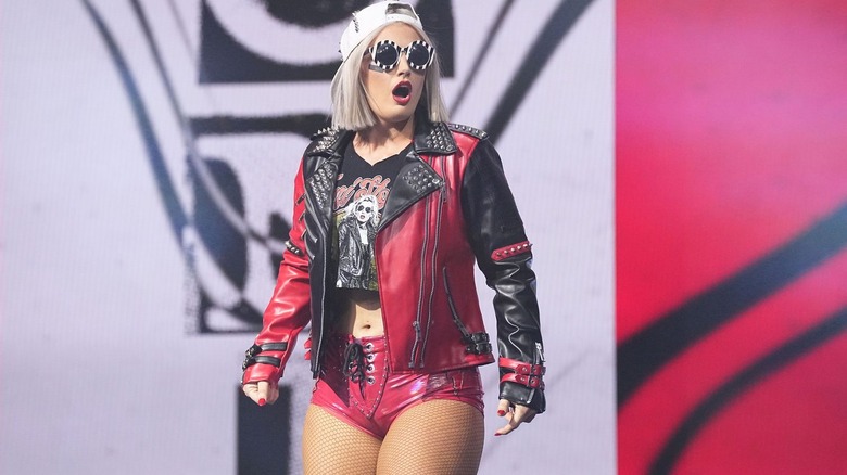 Toni Storm makes her entrance, donning a set of red and black gear, a leather jacket, and circular black sunglasses.