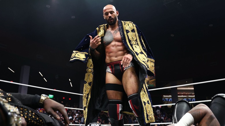 Ricochet dons a navy and gold coat as he stands above a downed Prince Nana and Swerve Strickland.