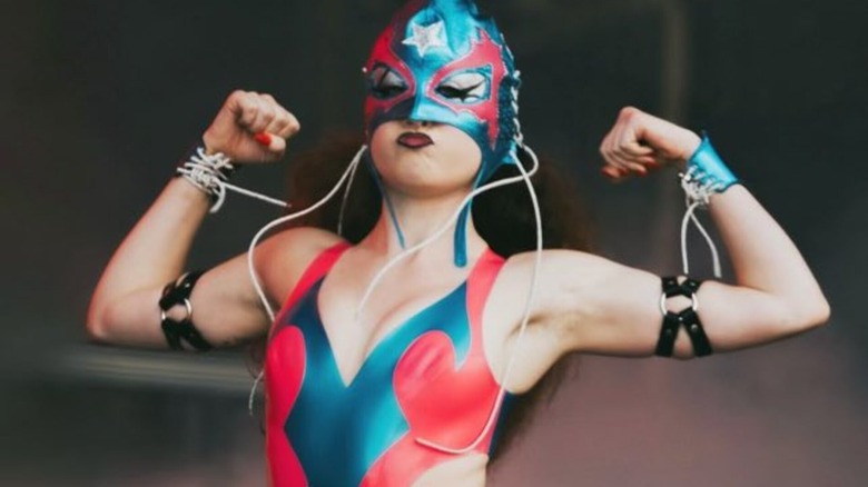 Chappell Roan flexes her biceps while sporting a blue, pink, and white lucha libre mask.