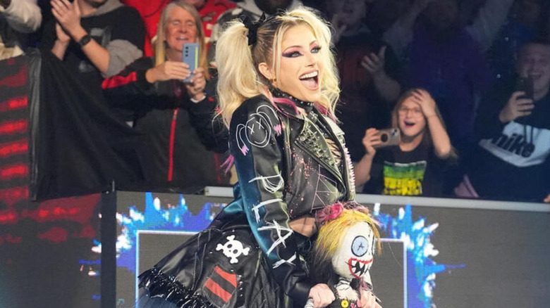 Alexa Bliss smiles as she appears at Royal Rumble 2025, dressed in a Play/Pain leather jacket, Gothic skirt, and a Lily doll.