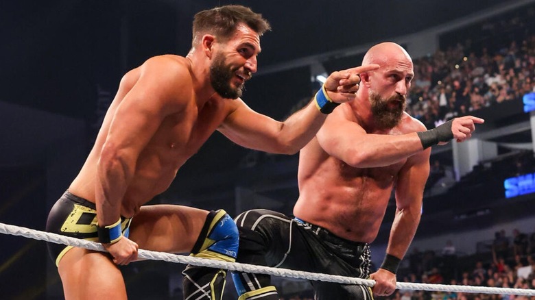 Johnny Gargano and Tommaso Ciampa stand on the ring ropes, smiling and pointing at Grayson Waller and Austin Theory on commentary during an episode of "WWE SmackDown."