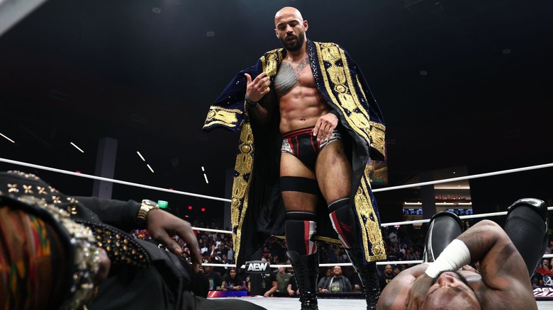 Ricochet dons a navy and gold coat as he stands above a downed Prince Nana and Swerve Strickland.