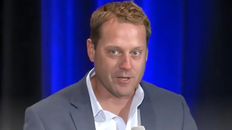 Former ESPN executive and current WWE head of Media and Production, Lee Fitting, sits on stage and addresses a crowd in a screenshot taken from a YouTube video.