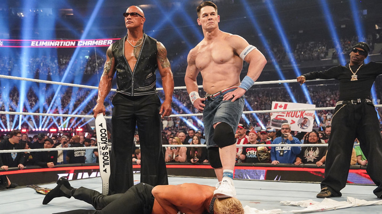 John Cena and The Rock stand over Cody Rhodes, while Travis Scott watches