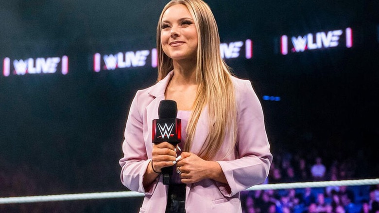 "WWE NXT" backstage interviewer Kelly Kincaid stands in the ring smiling, with a microphone in her hands.