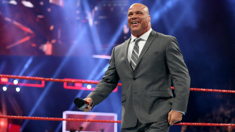 Kurt Angles smiles widely as he wears a neat suit and holds a microphone.