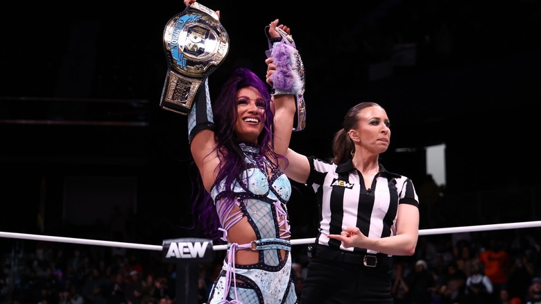 Mercedes Moné holds up the AEW TBS Championship and the NJPW STRONG Women's Championship as her hand is raised by referee Aubrey Edwards.