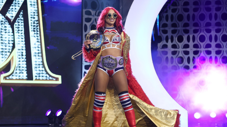 Mercedes Mone stands on the stage at an AEW event, holding her AEW TBS Championship and wearing the NJPW STRONG Women's Championship around her waist before a match.