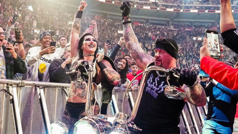 Women's World Champion Rhea Ripley stands beside The Undertaker, both of their arms raised in victory, with Undertaker riding a motorcycle, on 