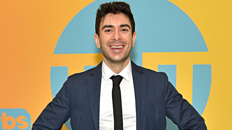 AEW President Tony Khan stands in front of a TNT sign during a red carpet event, smiling for the cameras.