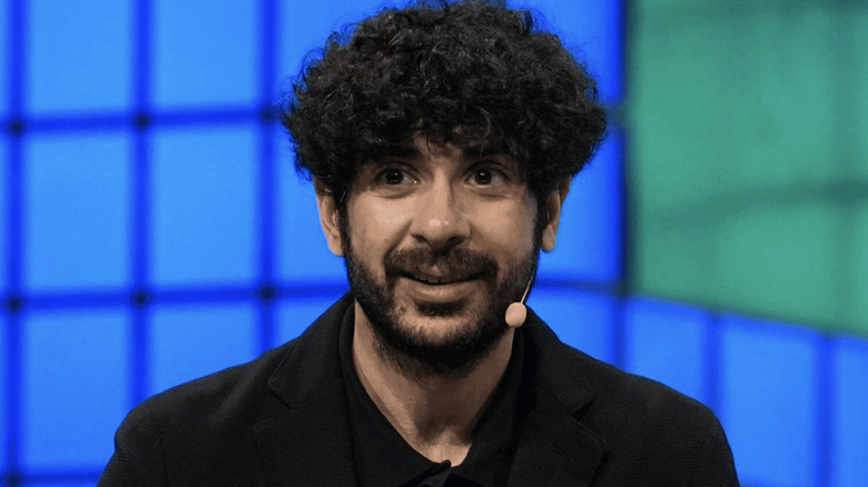 Tony Khan smiles in a black suit against a blue background, with a hands-free microphone taped to the right side of his face.