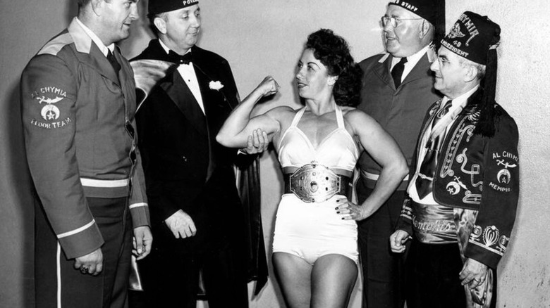 Mildred Burke poses wearing a championship belt for a group of men, one who is feeling her right bicep as she flexes.