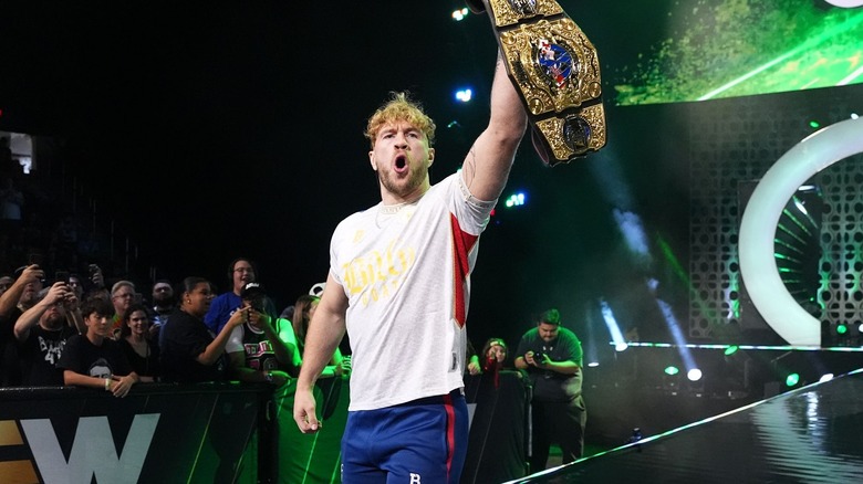 Will Ospreay brandishes the AEW International Championship as he comes down to the ring, dressed plainly in a white and red T-shirt and blue and red sweatpants.