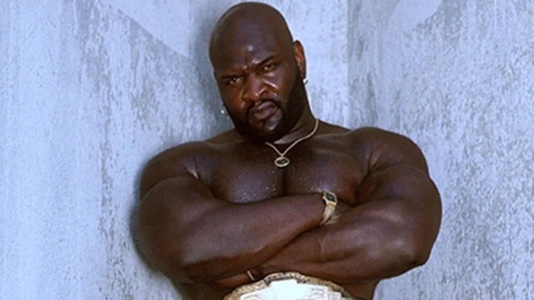 Ahmed Johnson posa frente a una pared con un cinturón de campeonato.