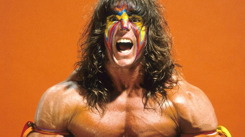 Ultimate Warrior poses backstage for a photoshoot, wearing his signature face paint an tassels on his arms before a match.