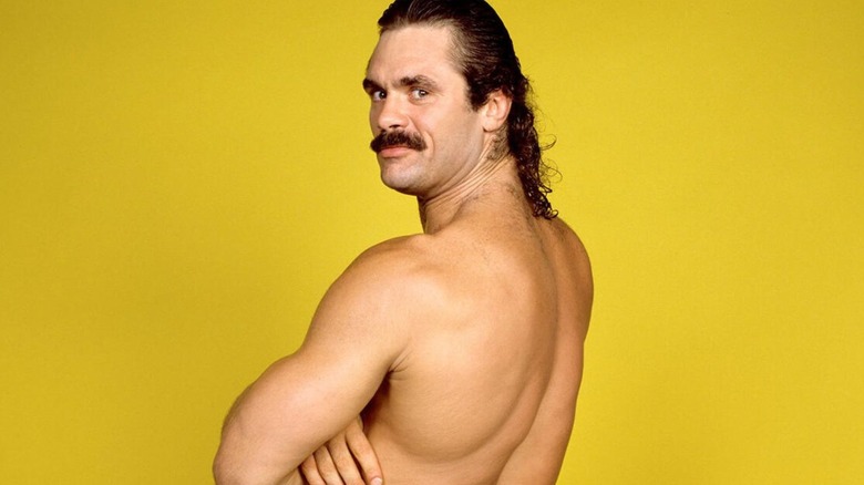 Rick Rude poses in front of a yellow backstage for a WWE photoshoot.