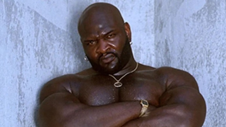 Ahmed Johnson poses for the camera, arms crossed, with a smirk on his face.