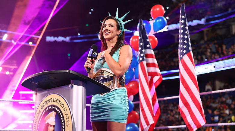 Chelsea Green, dressed as the Statue of Liberty, stands at a podium with a microphone in hand and the Women's United States Championship around her waist.