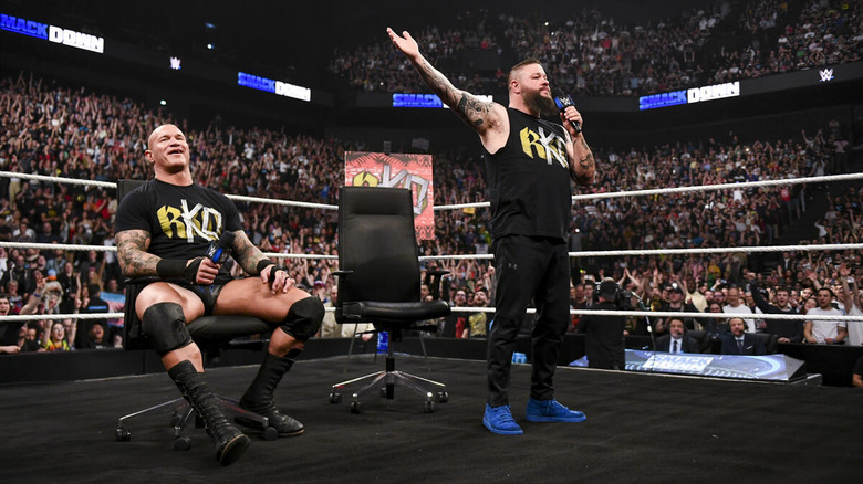 Randy Orton sits while Kevin Owens poses