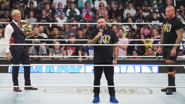 Kevin Owens speaks into microphone between Cody Rhodes and Randy Orton